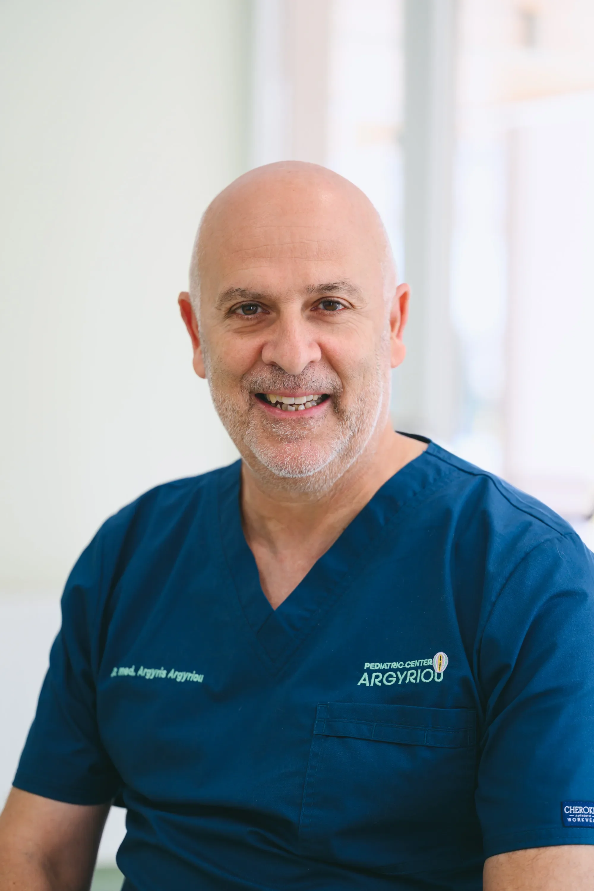 A smiling man doctor with a shaved head and gray beard wearing blue medical scrubs in a clinical setting.