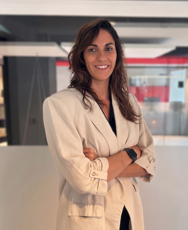 Mujer en un ambiente de oficina con cabello castaño y labios sonriendo, vestida con un blazer beige y con los brazos cruzados.