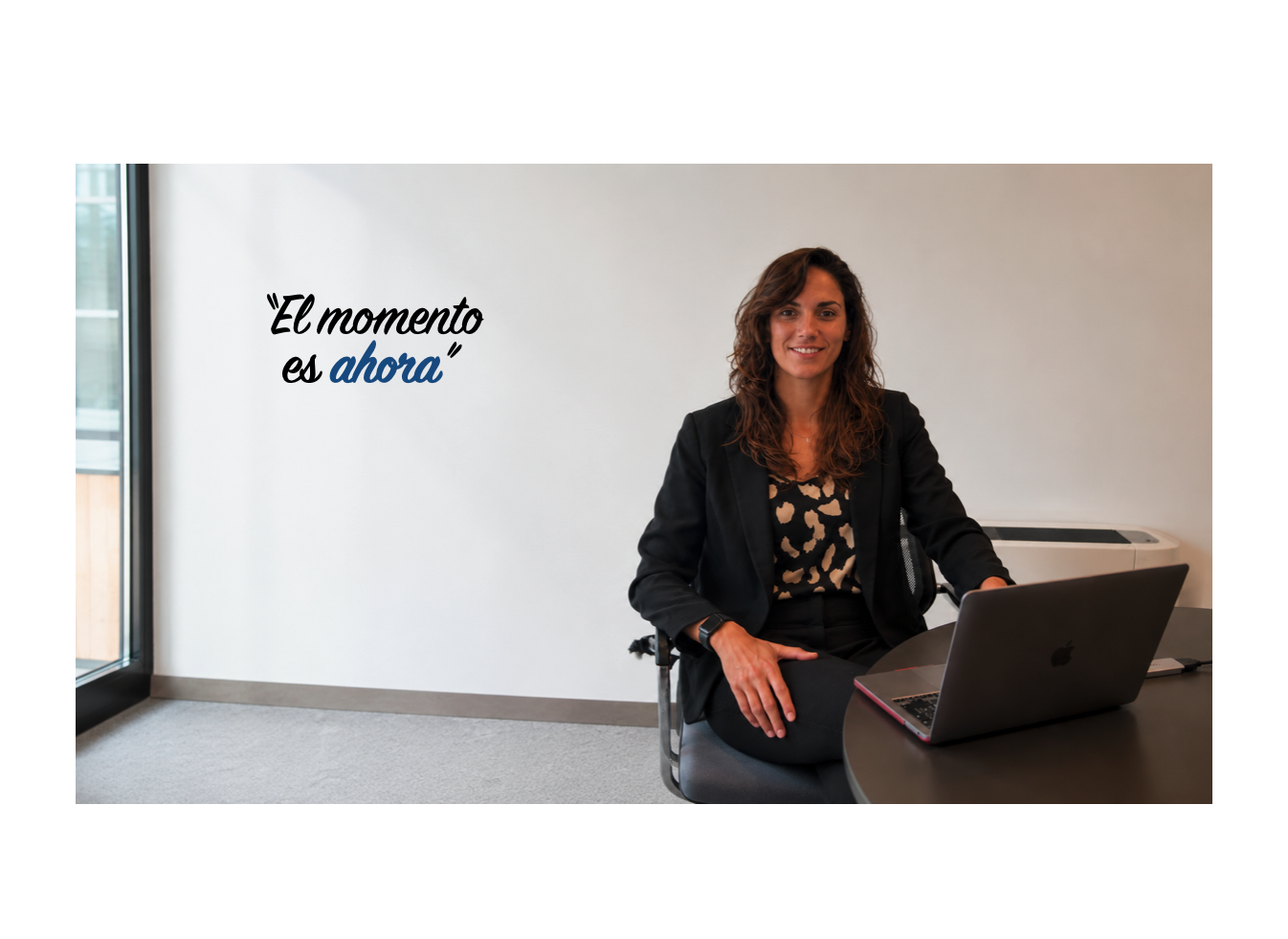 Mujer sonriente en una oficina, sentada en una silla junto a una mesa con una laptop y un teléfono, pared blanca con cita motivacional en letra cursiva que dice 'El momento es ahora'.