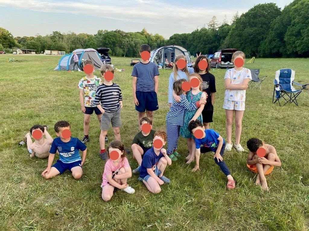 Group of kids at a campsite with tents and trees in the background, some sitting or kneeling on grass, others standing, posing for a photo.