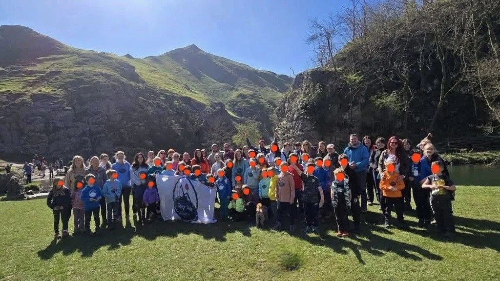 A large group of people, including children and adults, gathered outdoors on grassy terrain with mountains and a small lake in the background. The group is smiling and posing for a photo, some holding a flag with a bird emblem and other items.