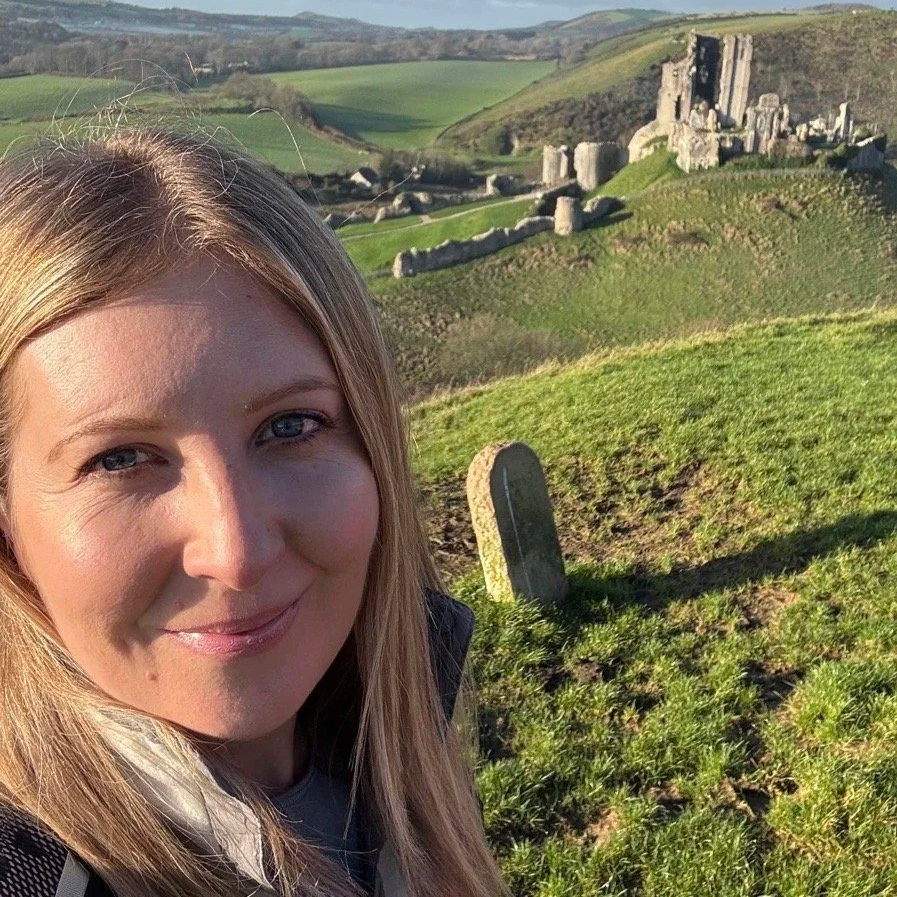A smiling woman with blonde hair taking a selfie in a lush green landscape with ancient stone ruins in the background.