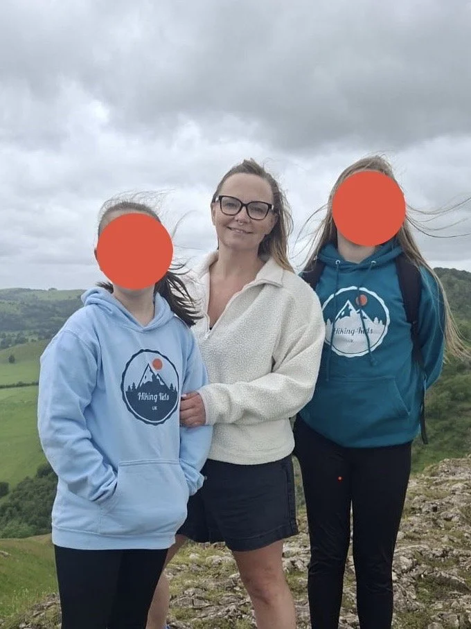 Three people standing outdoors on a hilltop with a cloudy sky in the background. Two young girls with hoodies featuring a mountain and sun logo, and an adult woman in a light-colored sweater. The girls' faces are obscured with orange circles.