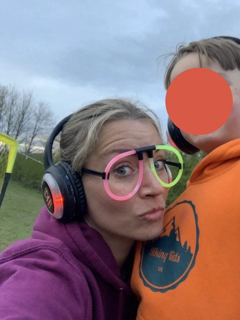 A woman with blonde hair wearing headphones and glasses with pink and green frames, making a duck face, standing next to a person wearing an orange hoodie with their face covered by an orange circle, outdoors during the daytime with a cloudy sky and trees in the background.