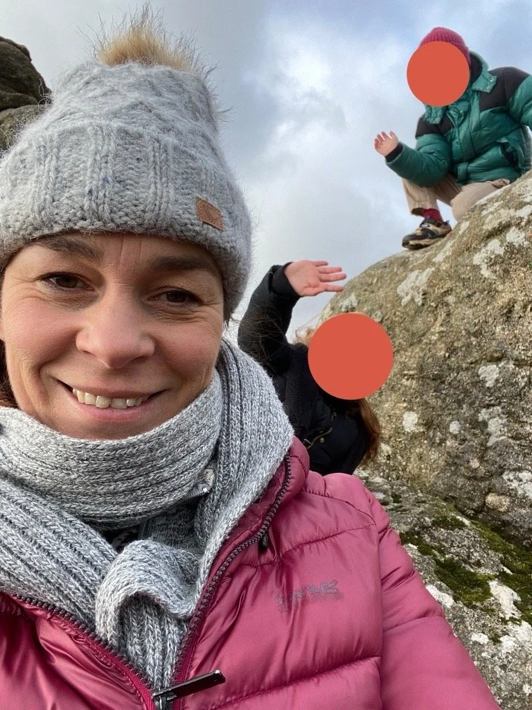 A woman wearing a gray knit beanie, gray scarf, and pink puffy jacket taking a selfie outdoors. Two children are climbing on a large rock behind her, with their faces covered by orange circles. One child is in a blue jacket and purple hat, and the other in black clothing with long hair.