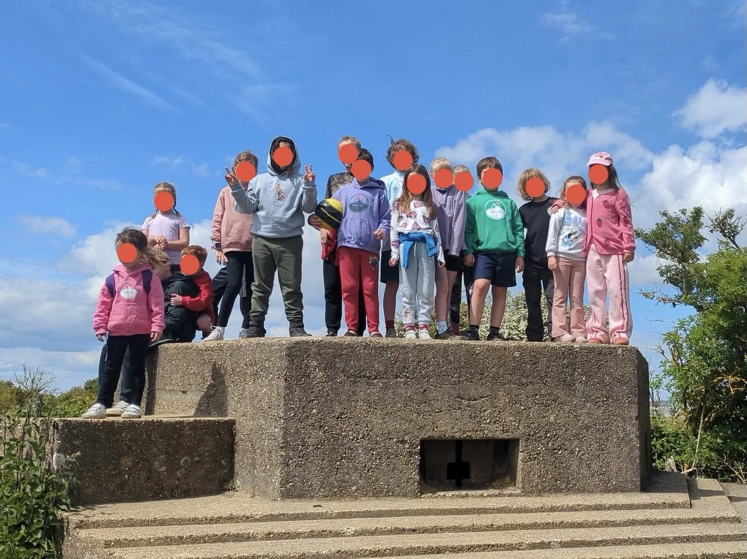 Group of children standing and sitting on a concrete monument outdoors against a blue sky with some clouds, some making peace signs, dressed in casual clothes and backpacks.