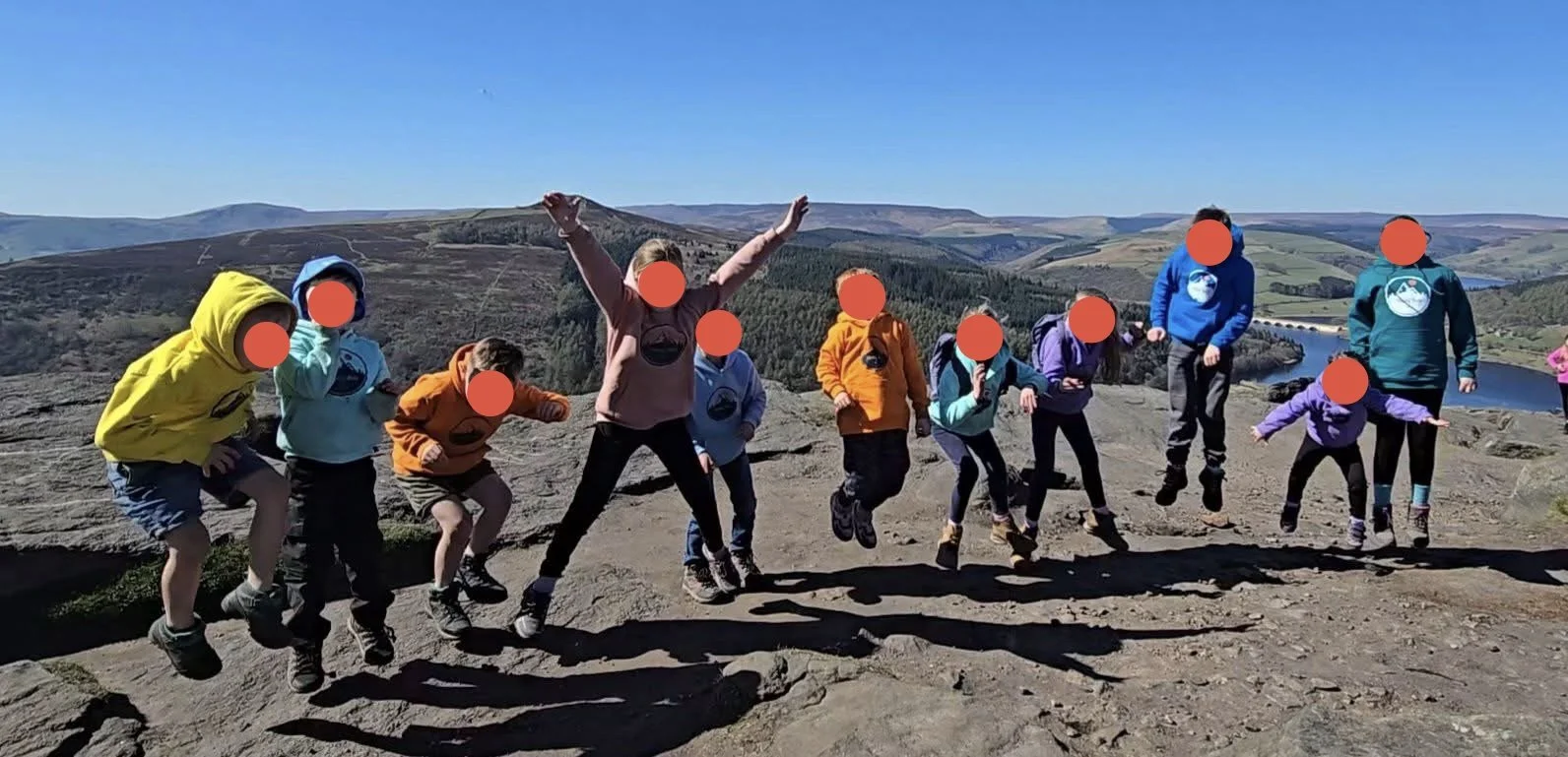 Group of children jumping on a rocky hilltop with a scenic view of hills, a river, and a clear blue sky in the background.