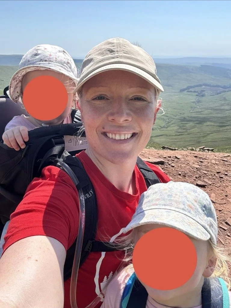 A woman, smiling, wearing a beige cap and red shirt, hiking outdoors on a trail with two young children, both wearing hats, in a scenic green landscape with rolling hills in the background.