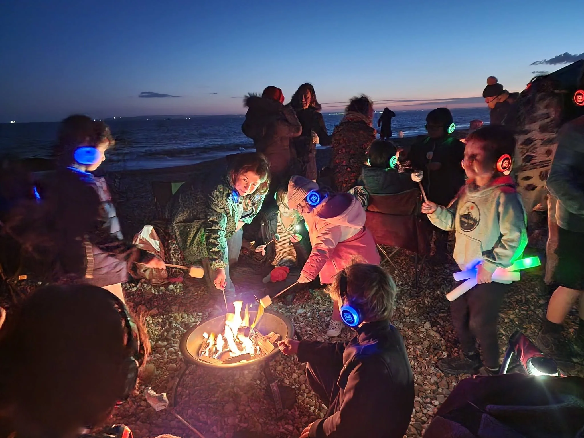 A group of children and adults gathered around a campfire on a rocky beach at dusk, some wearing colorful headphones and holding sticks with marshmallows, with the ocean and a darkening sky in the background.
