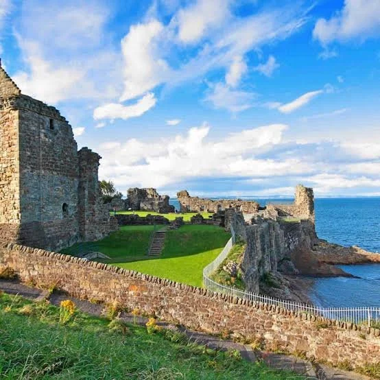 Ruins of an old stone castle on a grassy hill overlooking the ocean with a bright blue sky and scattered clouds.