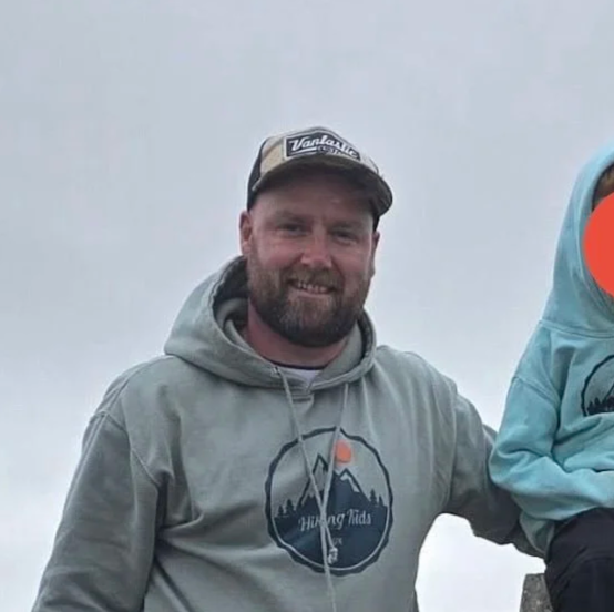 A bearded man in a hoodie and cap smiling outdoors, with part of a person in a blue jacket visible on the right.