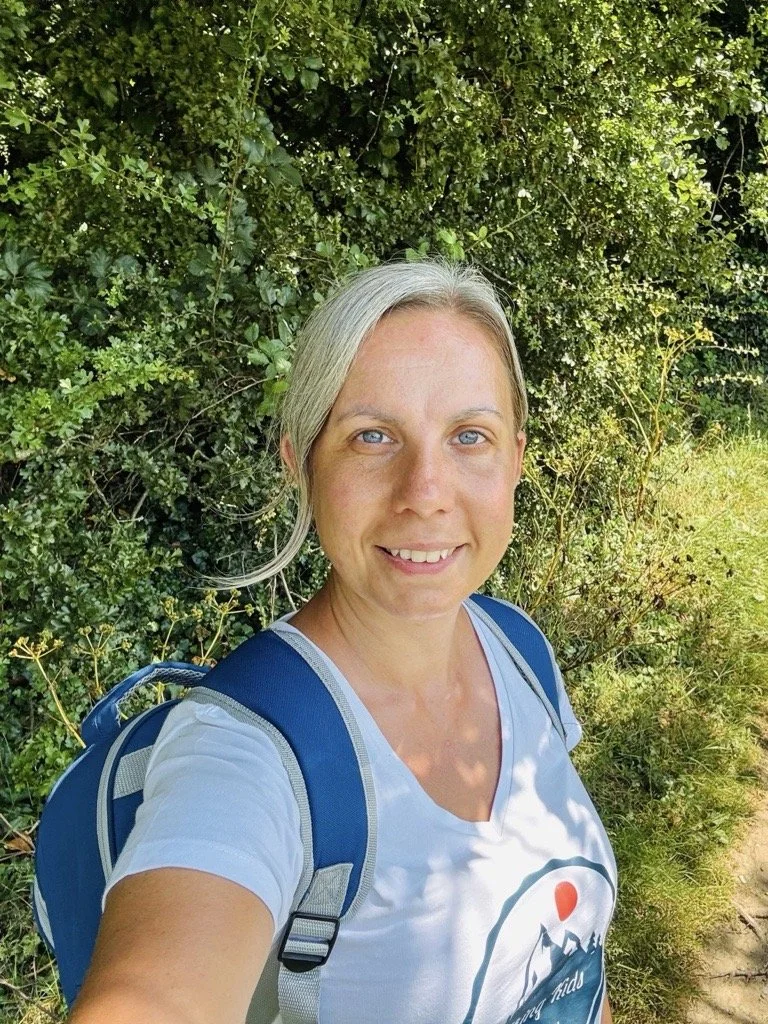 A woman with blonde hair, blue eyes, and a white t-shirt with a hiking logo, taking a selfie outdoors with green bushes and trees in the background.