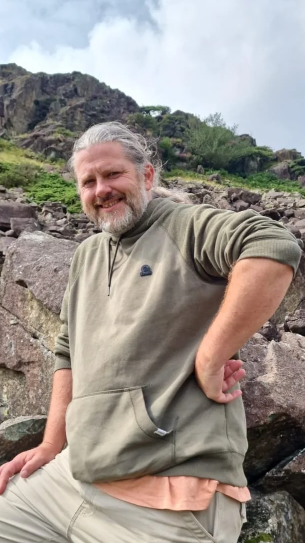 A man with long gray hair and a beard posing outdoors in a rocky, mountainous area with green vegetation and a partly cloudy sky.