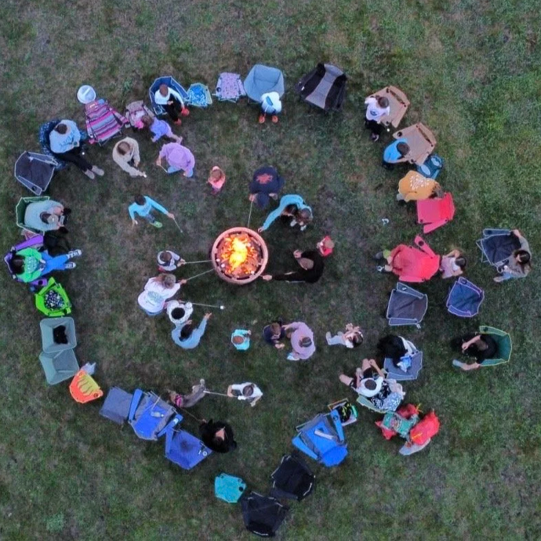 People gathered around a campfire in a circle on a grassy field, with camping chairs and tents nearby.