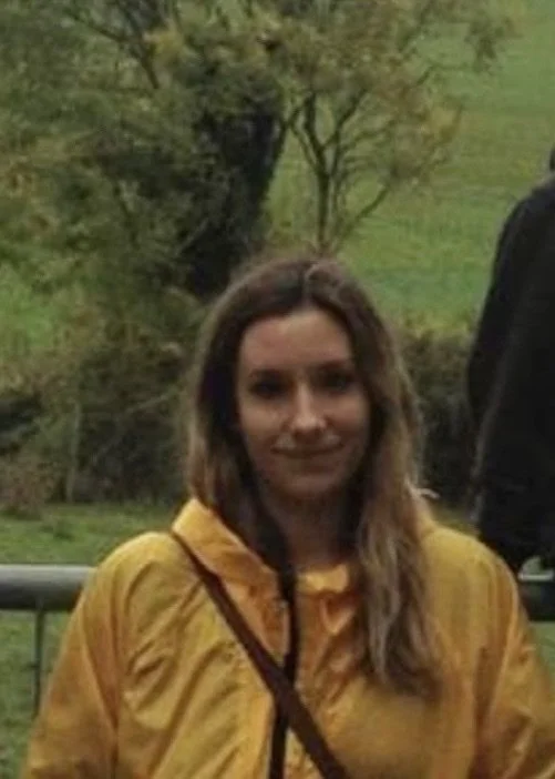 A woman with long brown hair, wearing a yellow jacket, standing outdoors with trees and greenery in the background.