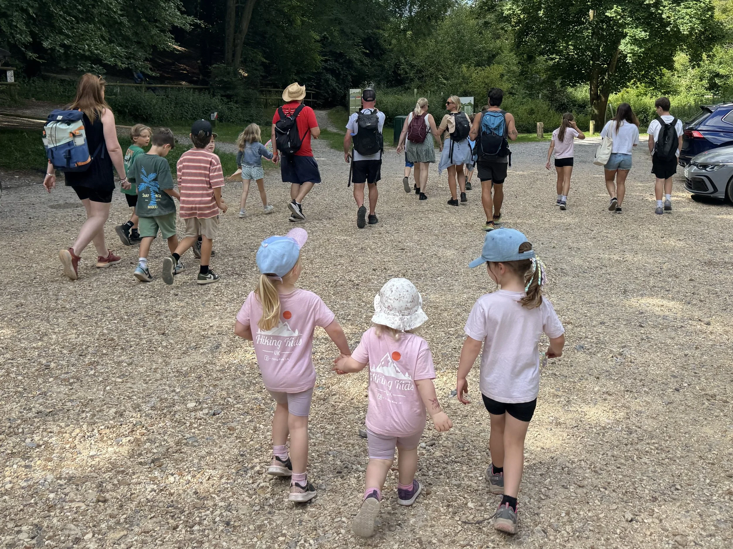 A group of children and adults walking together outdoors on a gravel path surrounded by green trees, some carrying backpacks and wearing casual clothing.