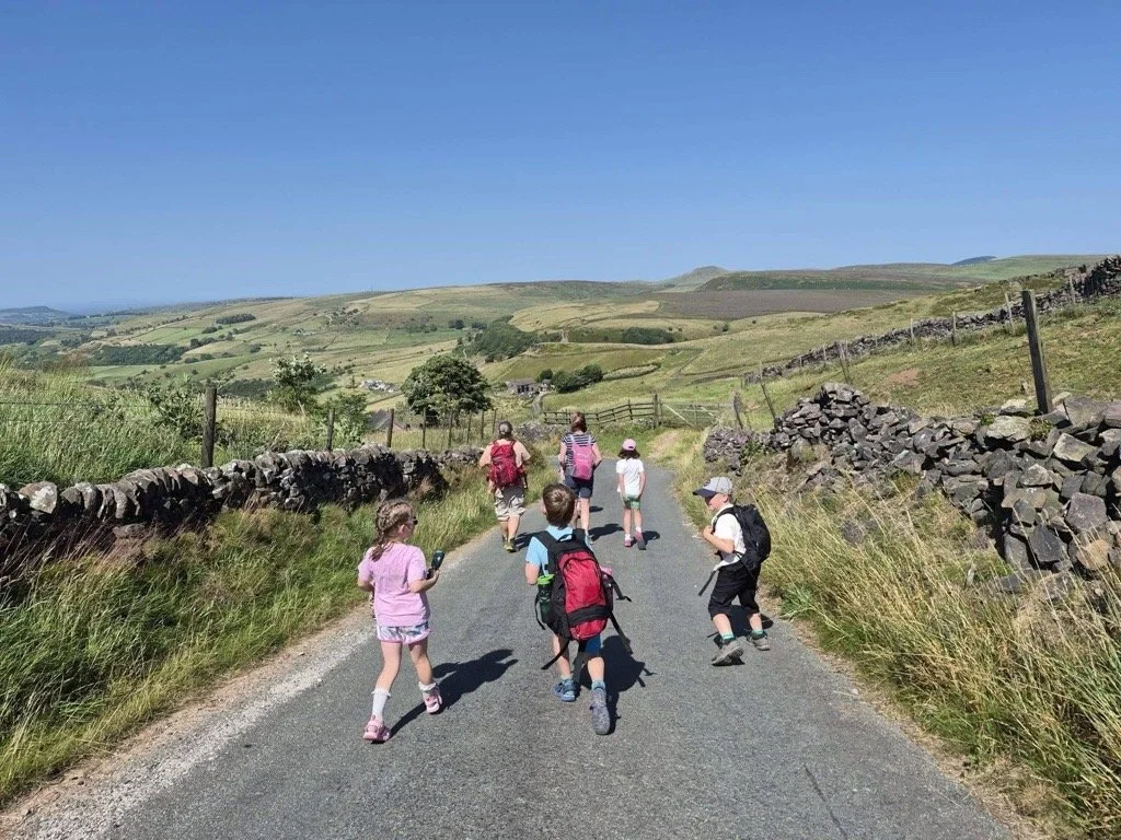 A group of children and adults walking on a rural country road surrounded by stone walls and green rolling hills under a clear blue sky.