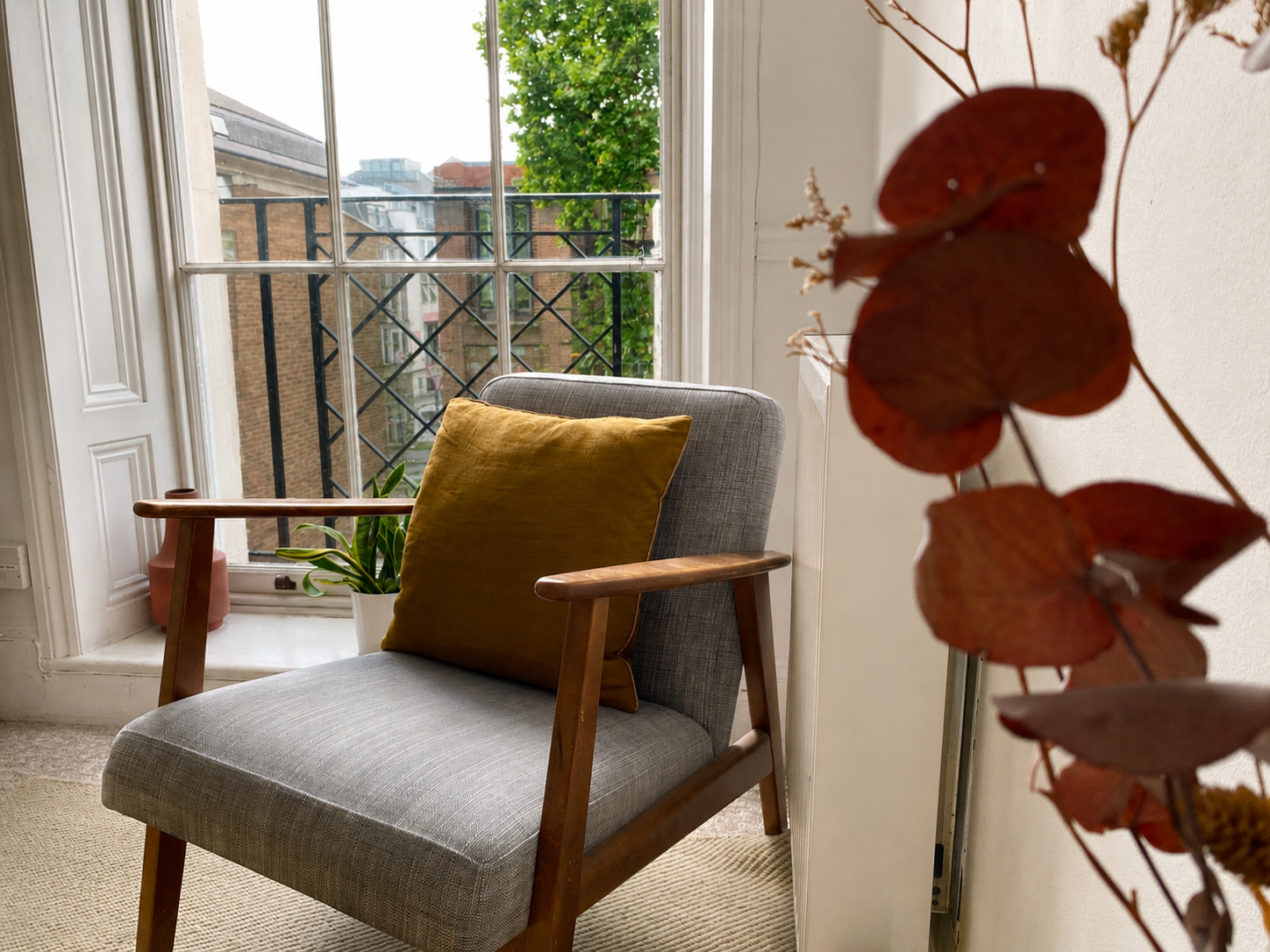An empty grey chair with a wooden frame and a yellow cushion in the corner of a brighttherapy room. In the foreground, framing the chair, is an out of focus red flower. Behind the chair, a large window revealing buildings and trees across the street.