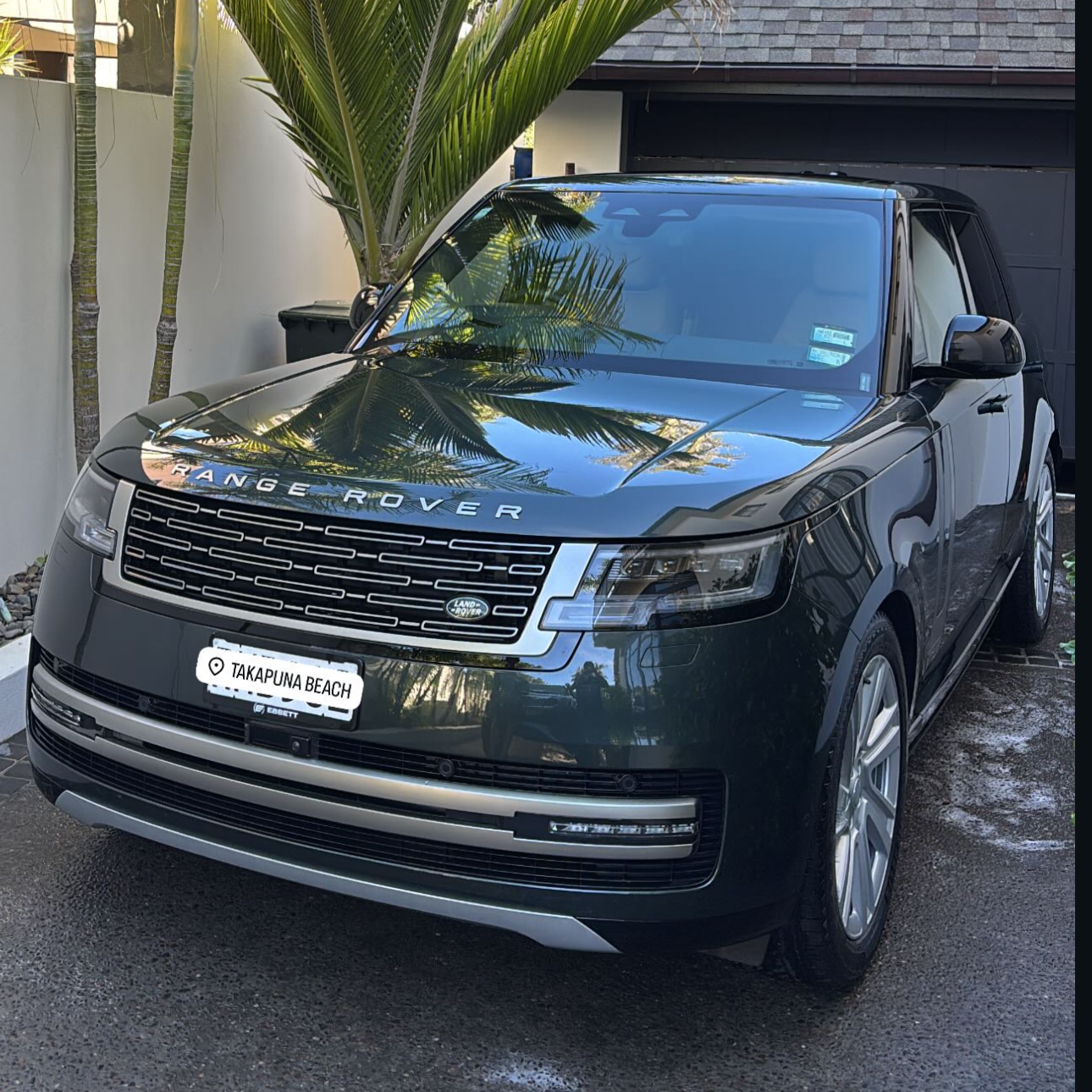 Black Range Rover parked in driveway with a palm tree and garage in the background, reflecting the palm tree and sky on its hood and windshield, with a beach location label 'Takapuna Beach' on the front license plate.