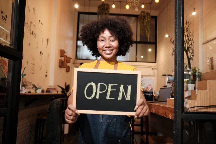 A woman with curly hair smiling and holding a chalkboard sign that says 'OPEN' inside a cozy cafe or shop.