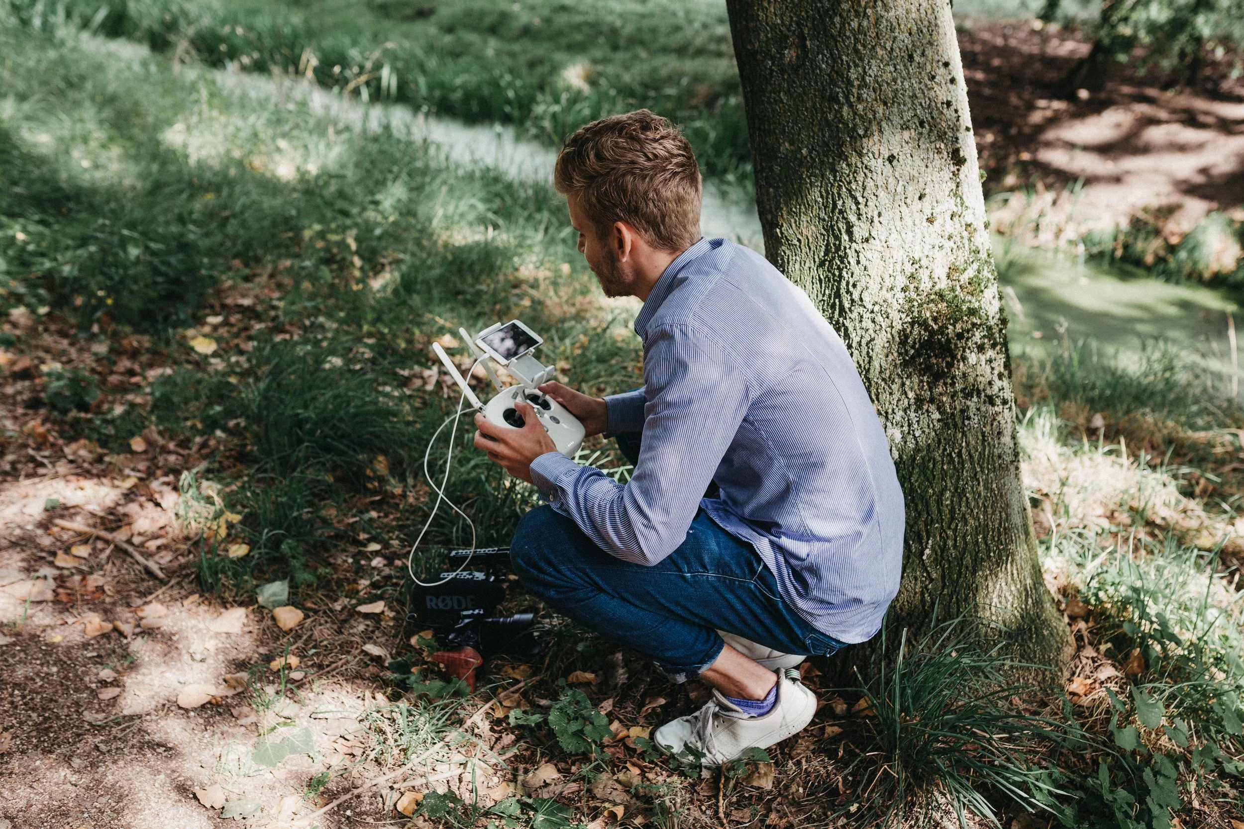 A man kneeling on the ground next to a tree outdoors, operating a drone controller with an aerial drone nearby. He is focused on the drone controller, wearing a light blue striped shirt and dark jeans.