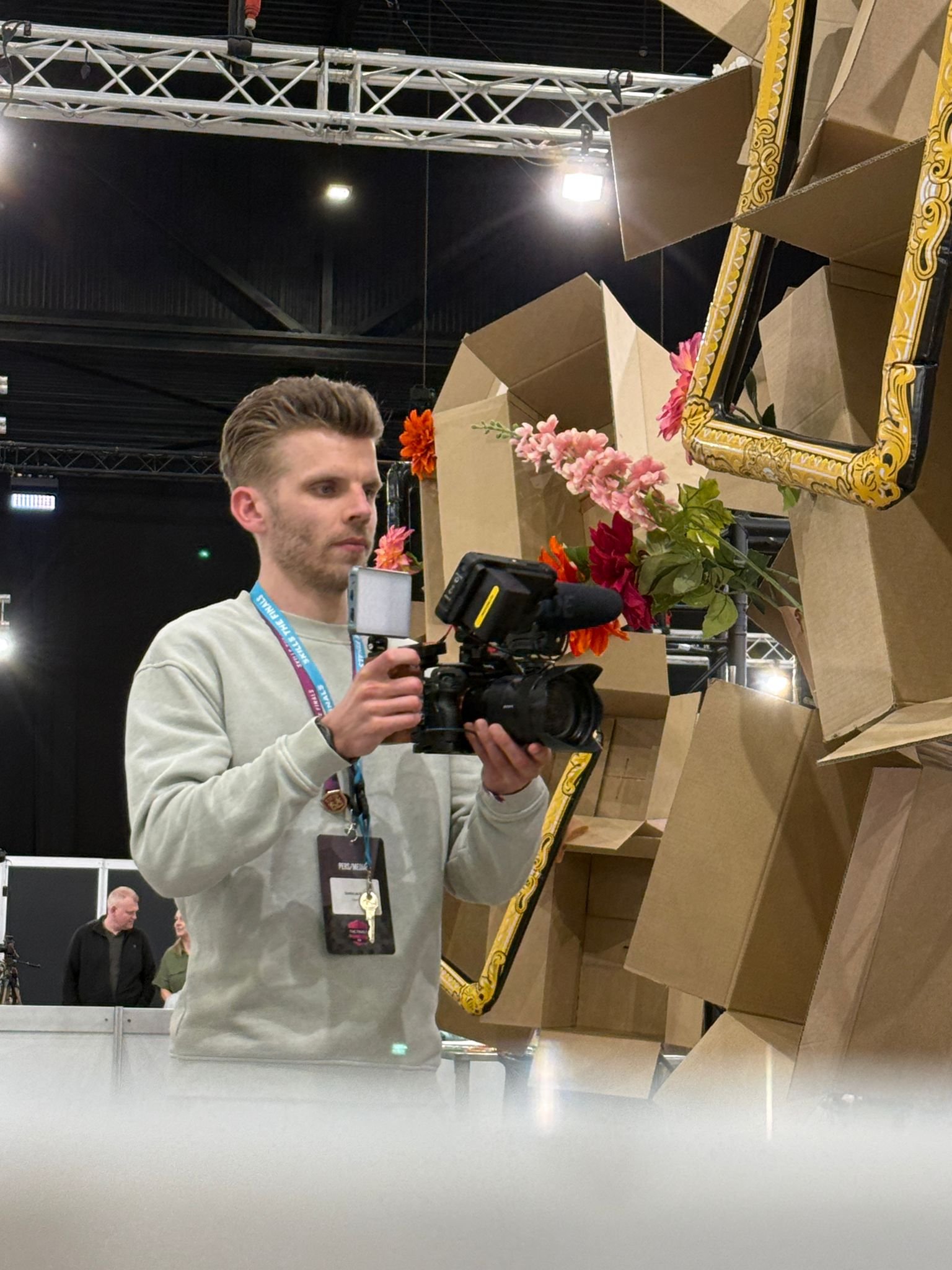A young man with light brown hair and a beard is holding a camera and looking at a mirror while taking a photo. The mirror reflects cardboard boxes, pink and orange flowers, and part of the exhibition area with black ceiling panels and lighting.