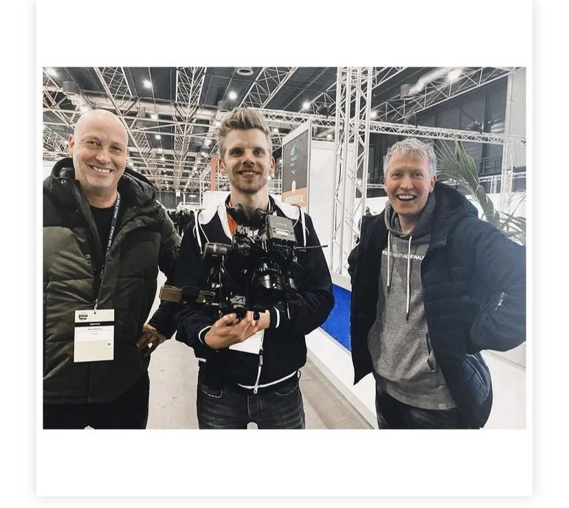 Three smiling men standing at a trade show or conference, with one holding a professional camera, surrounded by exhibition booths and metal framework overhead.