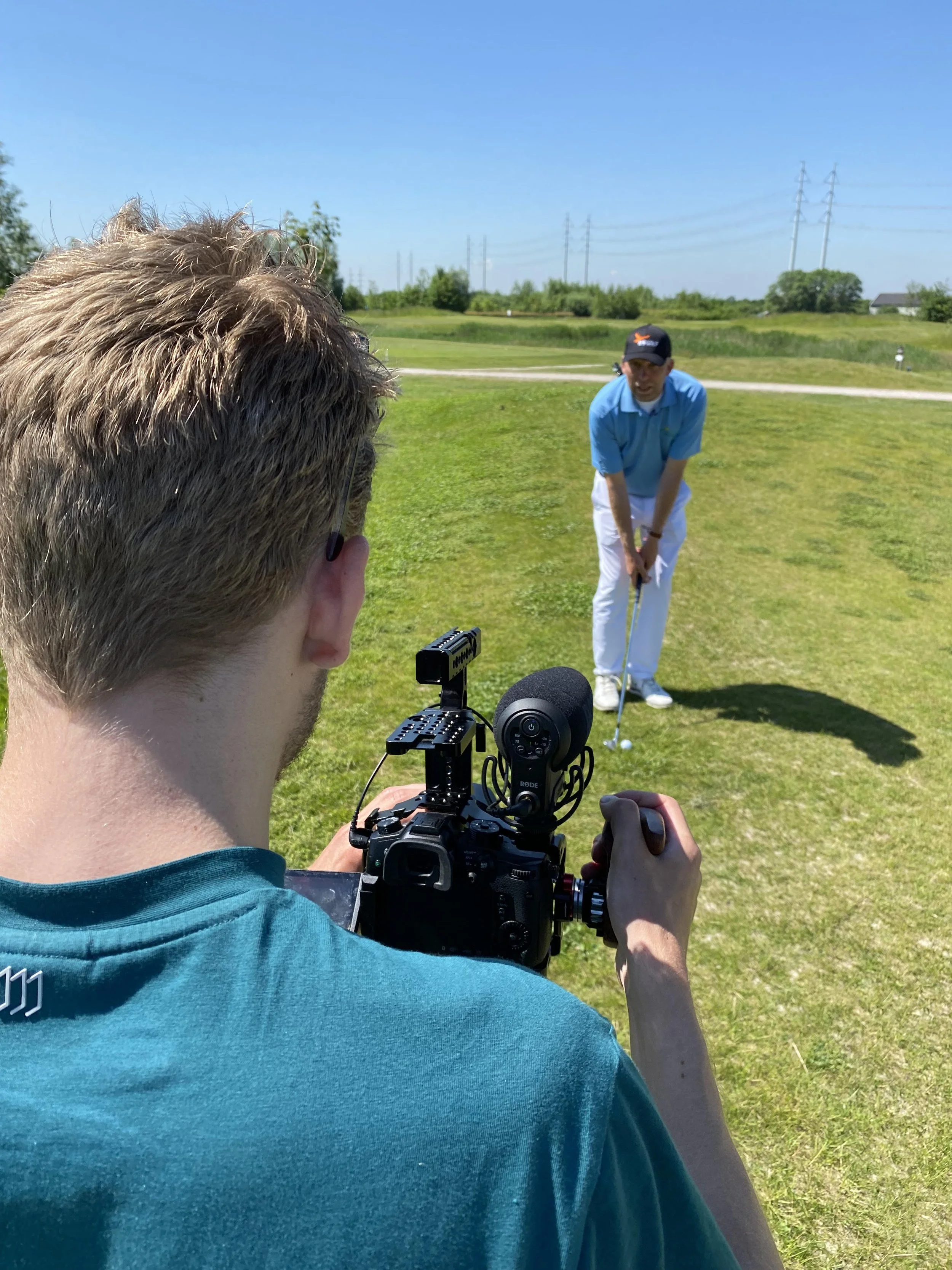 A person in a teal shirt is filming a golfer on a golf course using a camera mounted on a stabilizer. The golfer is wearing a blue shirt, white pants, and a black cap, preparing to hit a golf ball on the lush green fairway under a clear blue sky.