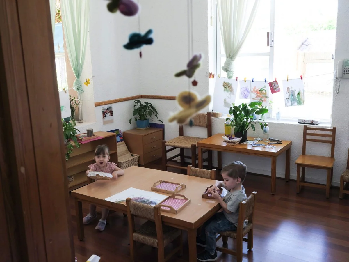 Dwoje małych dzieci, dziewczynka i chłopiec, siedzi przy drewnianym stole w nasłonecznionym pokoju o białych ścianach, rysując i bawiąc się zabawkami. W pokoju znajdują się duże okna, rośliny w doniczkach, dziecięce rysunki rozwieszone na sznurku oraz różne meble.