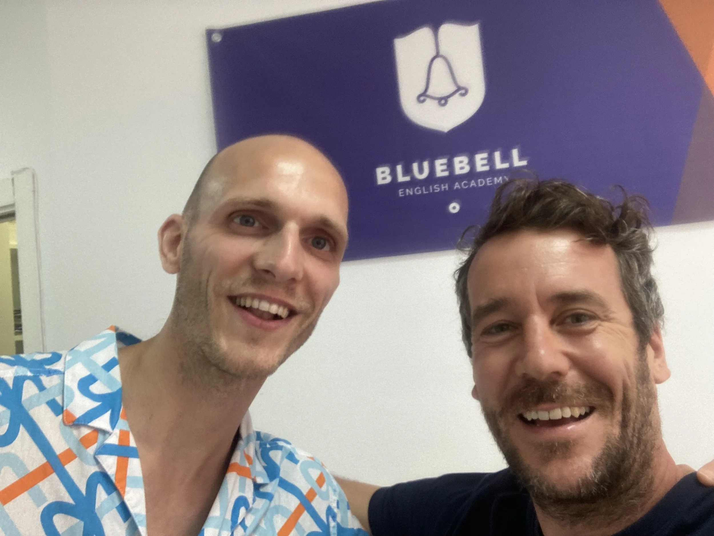 Two men are smiling and taking a selfie in front of a blue sign that reads "Bluebell English Academy" with a bell logo.
