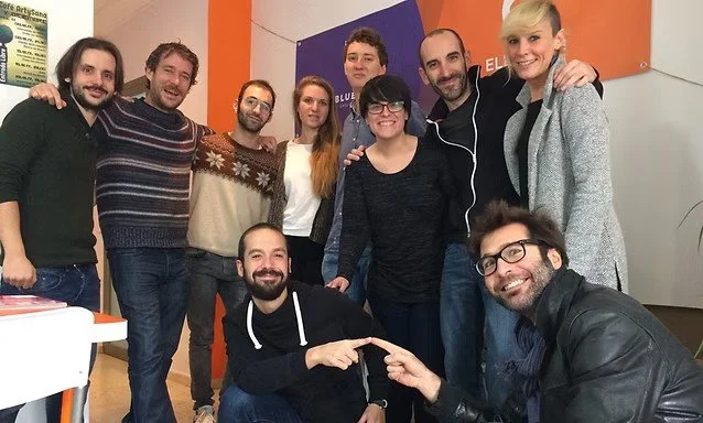 Group of nine people standing together indoors, smiling at the camera. One person kneeling in front, pointing at another person sitting on the ground. The background has colorful walls and posters.