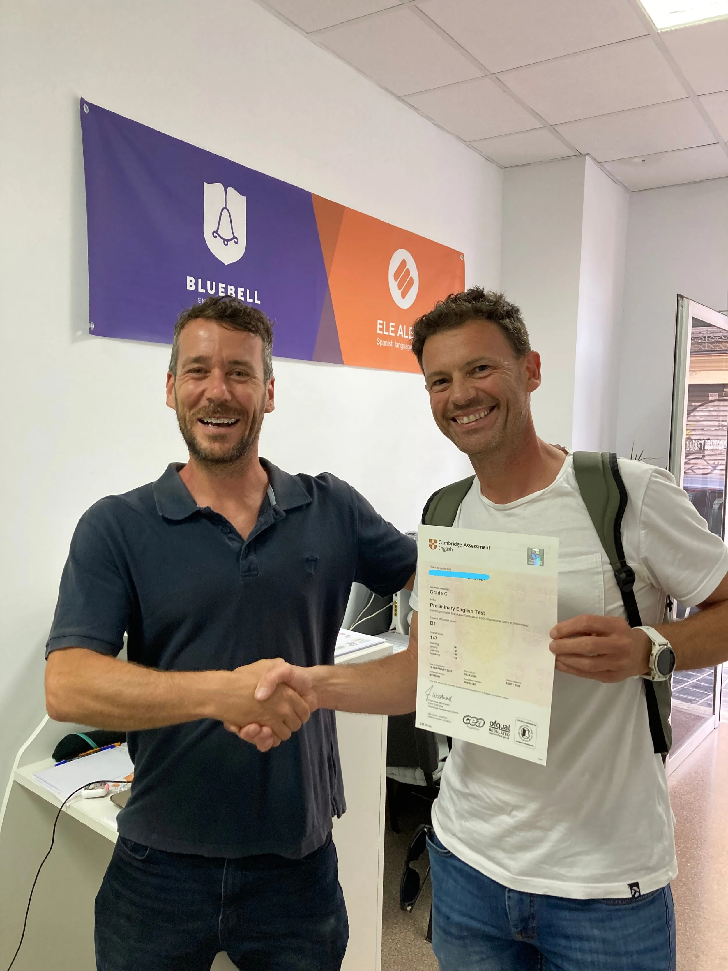 Two men shake hands in an office environment, smiling. One man holds a completed English proficiency test certificate. Behind them, a sign displays 'Bluebell' and 'Ele Al', indicating a language center or testing location.