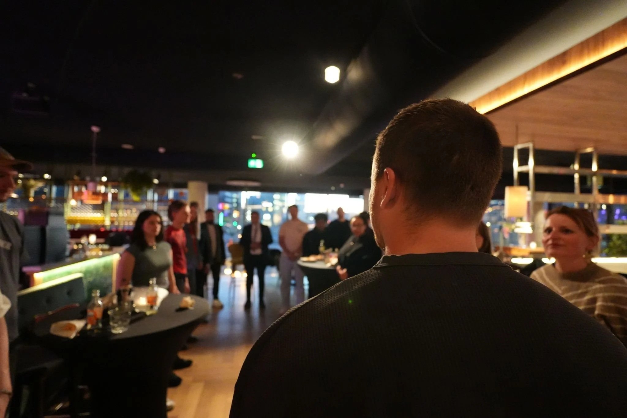 Een groep mensen staat en zit in een drukke, gezellige bar of restaurant met moderne verlichting en kleurrijke decoraties.