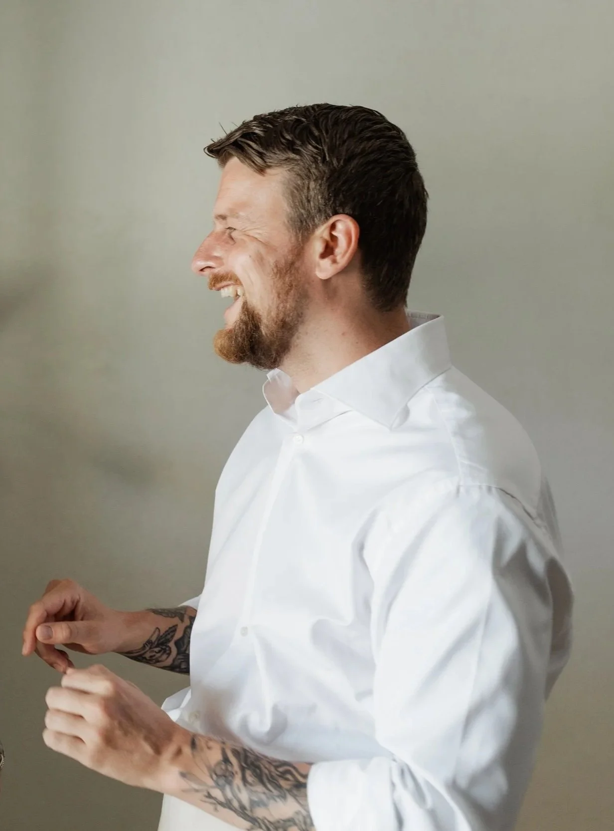 Een lachende man met een korte, donkere haarstijl en een gezicht met een lichte baard en snor, gekleed in een witte overhemd, die naar rechts kijkt, met tattoos op zijn onderarmen, tegen een effen grijze achtergrond.