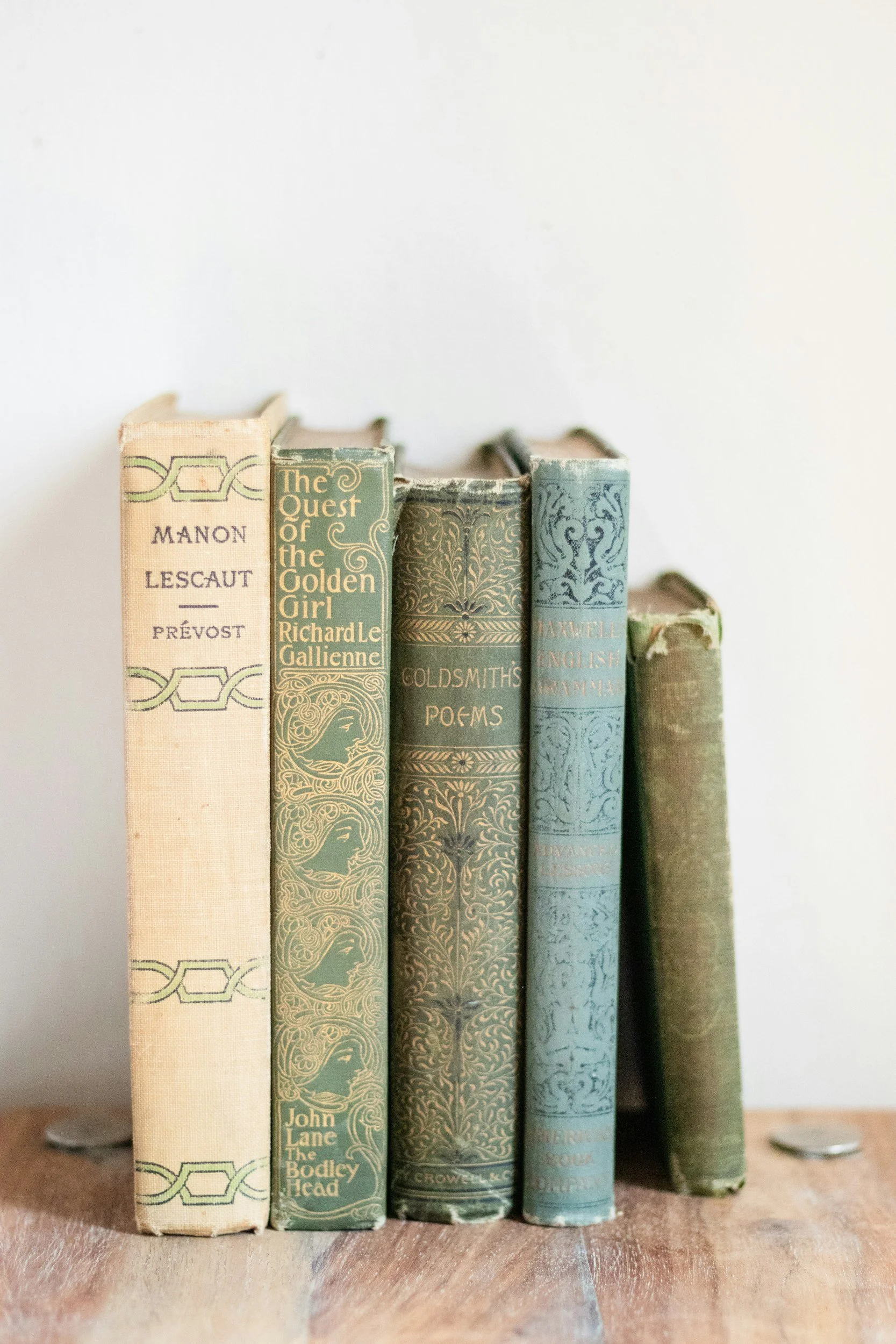 A row of vintage hardcover books with faded spines and ornate designs on a wooden surface, with a plain light-colored background.