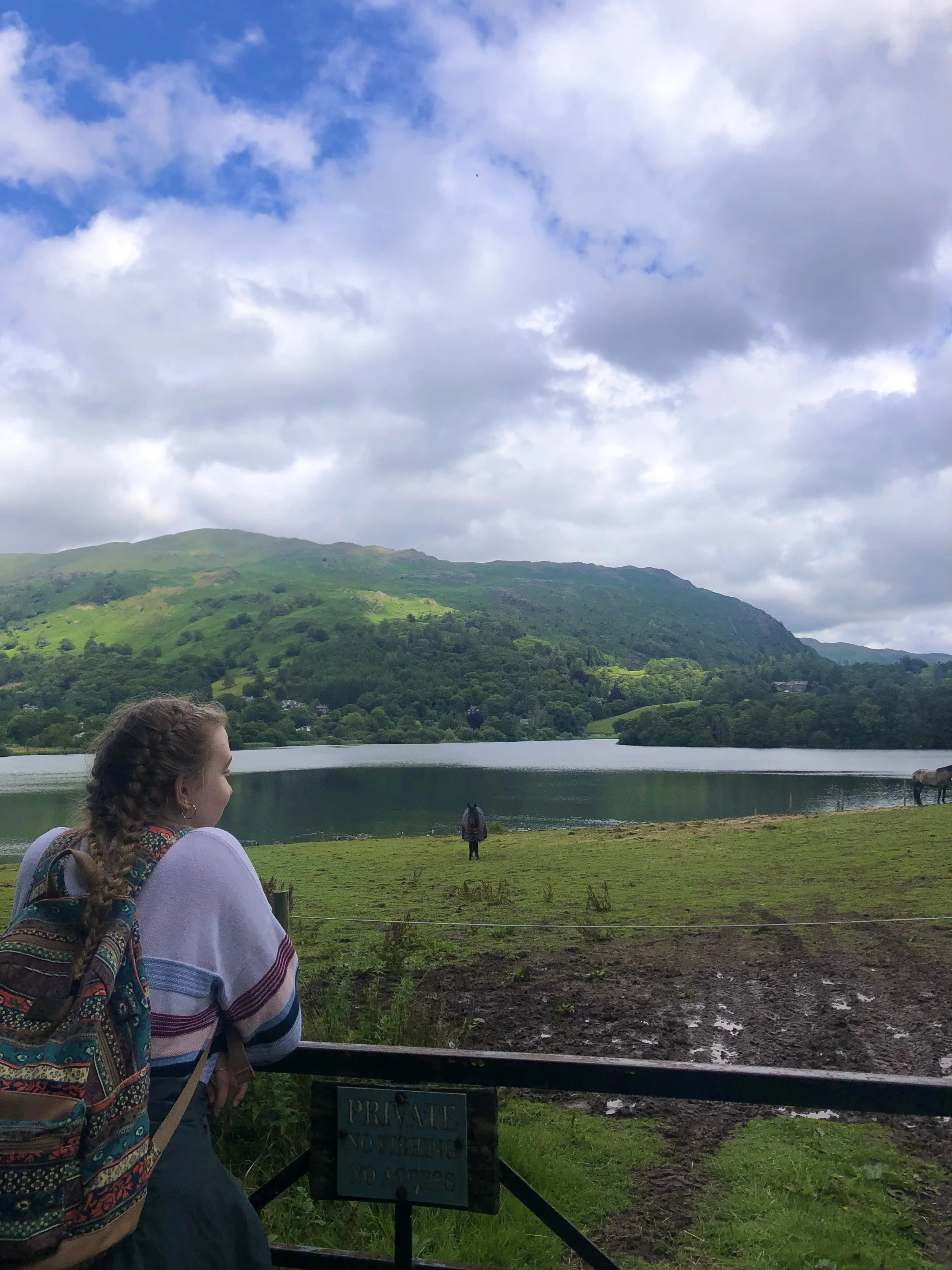 A woman with braided hair and a colorful backpack is sitting on a bench near a lake. In front of her, two people are standing near the water's edge. The lake is surrounded by lush green hills and mountains under a cloudy sky.