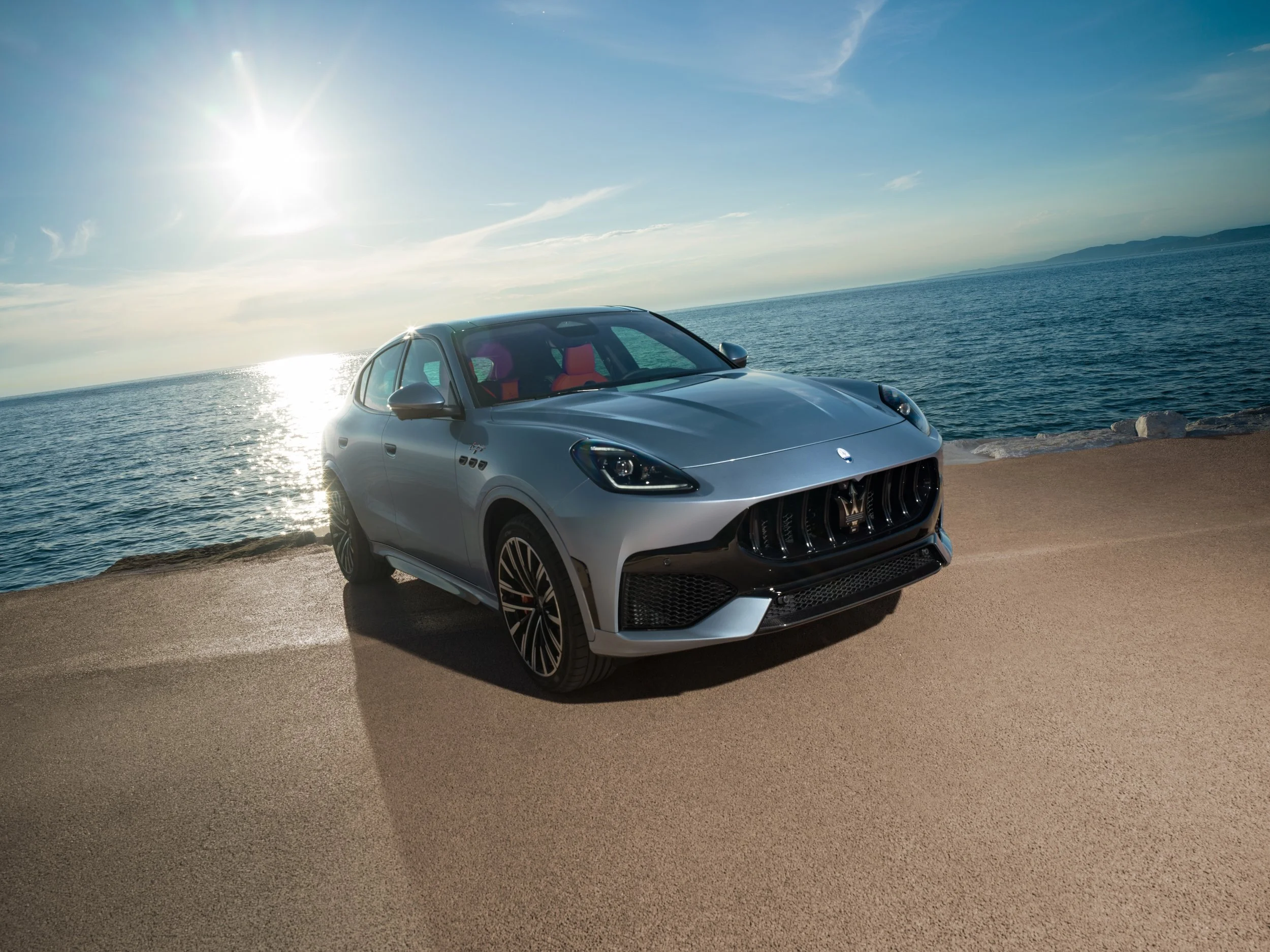 A silver Maserati SUV parked by a body of water with a rocky shoreline, under a clear blue sky with the sun shining brightly.