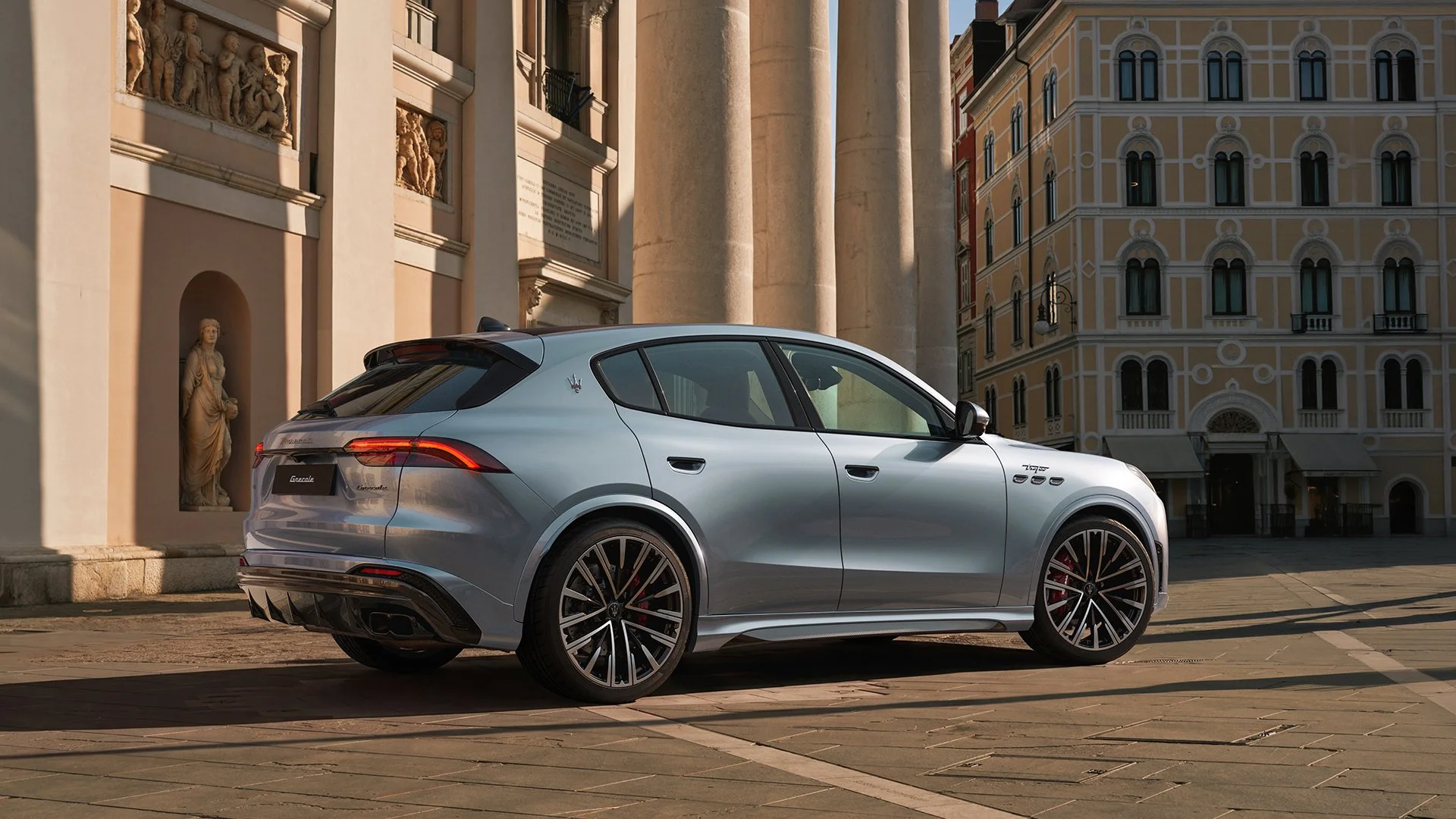 A silver Maserati Grecale parked in an urban setting with historic European architecture, including classical columns, sculptures, and ornate buildings in the background.