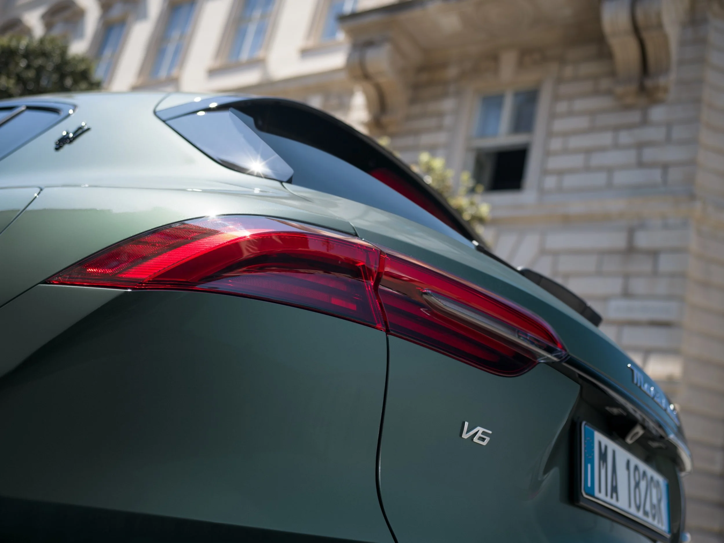 Close-up of the rear end of a green luxury car, focusing on the taillight, rear window, and badge, with part of a European license plate visible, parked in front of a building with classical architecture.