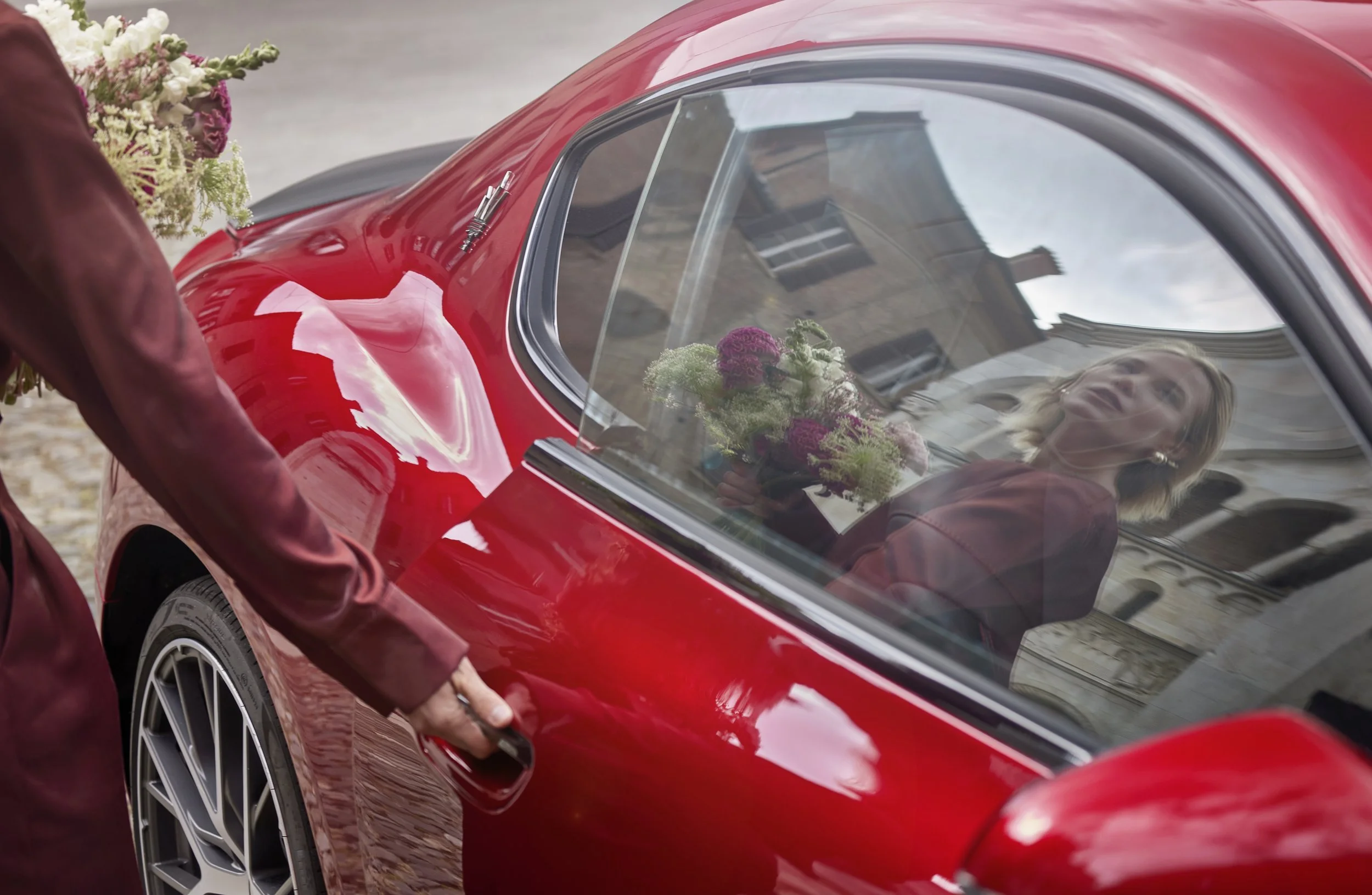 A woman is sitting inside a red sports car with a flower bouquet visible on the interior. A person is standing outside the car near the door, which is partially open. The scene appears to be outdoors in daylight.