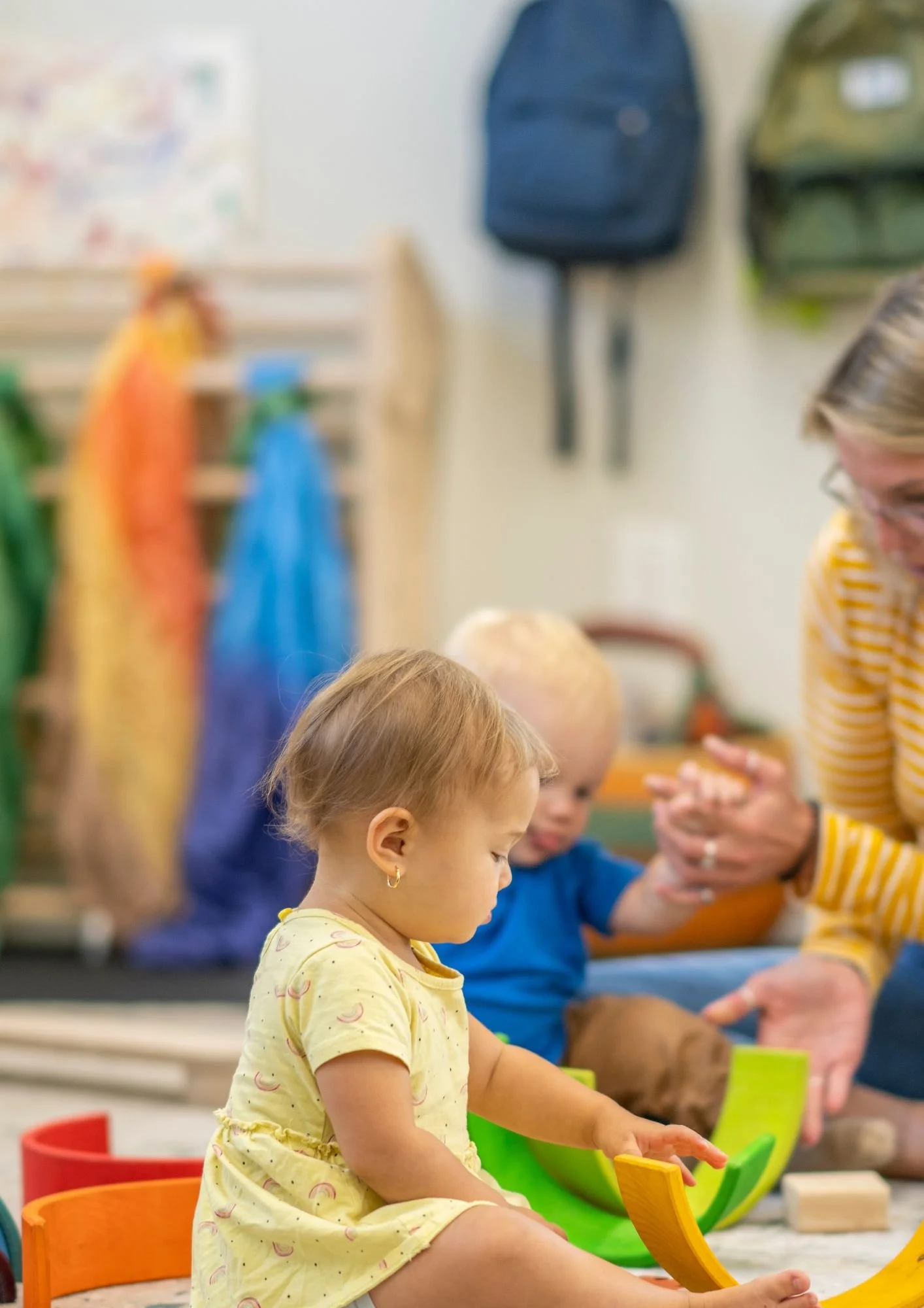 Toddler playing with colorful toys in a classroom while adults and other children sit nearby