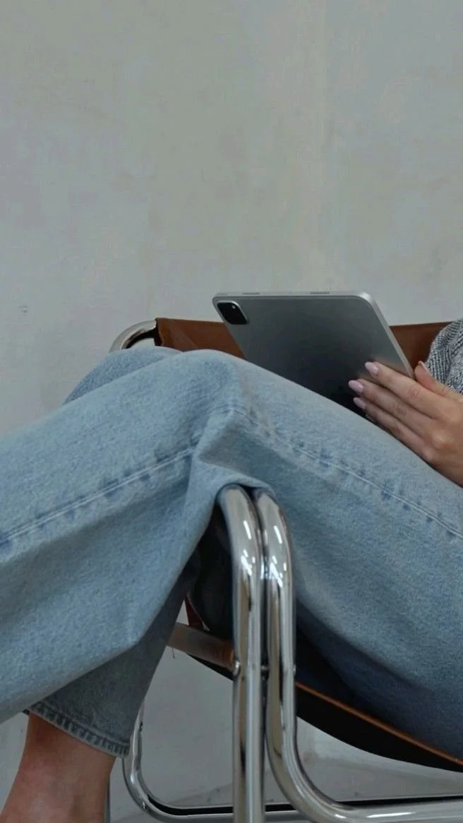 A person sitting in a chair, wearing jeans, holding a tablet in their hand. The person is in a plain room with a neutral wall.