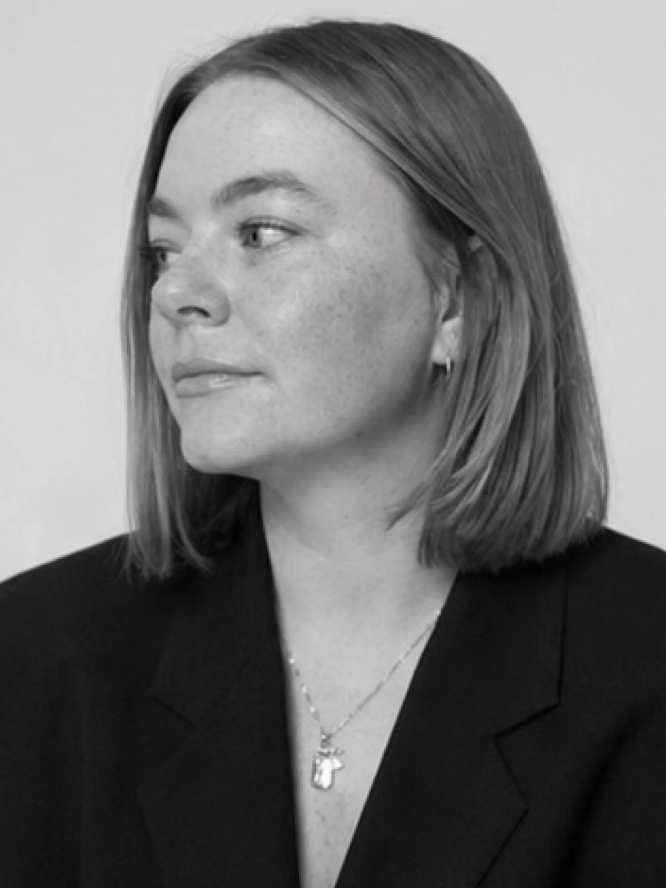 Black and white photo of a woman with straight hair, wearing a black blazer, a white top, and a necklace with a cross pendant.