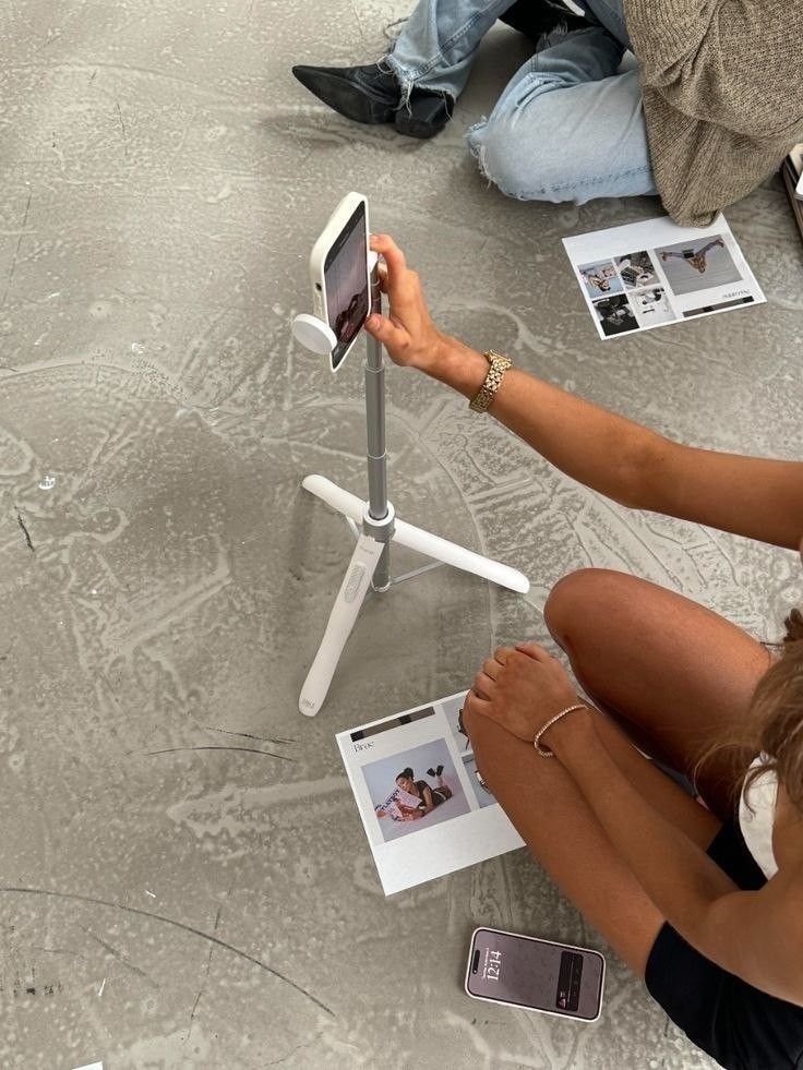 Person taking a photo with a smartphone mounted on a tripod, sitting on a concrete floor with printed photos nearby.
