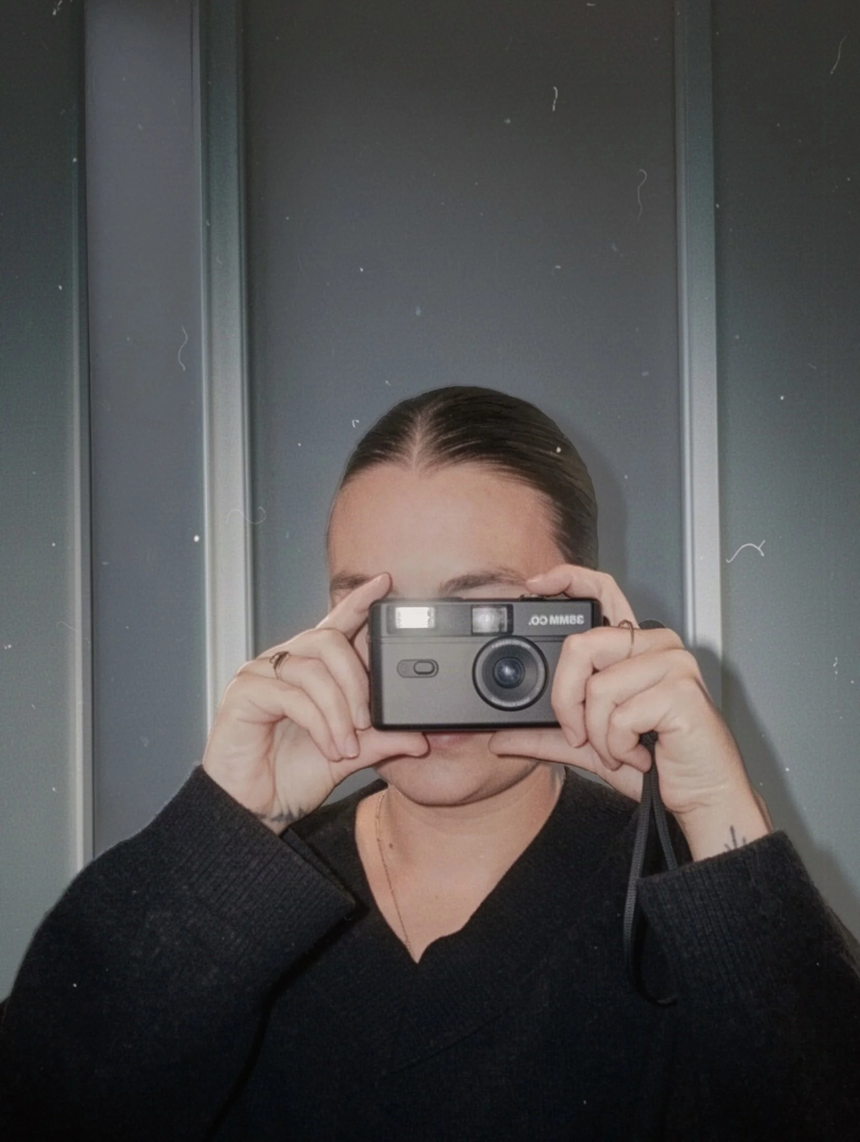 Person taking a mirror selfie with a vintage camera, wearing a black sweater, in a space with metallic panel walls.