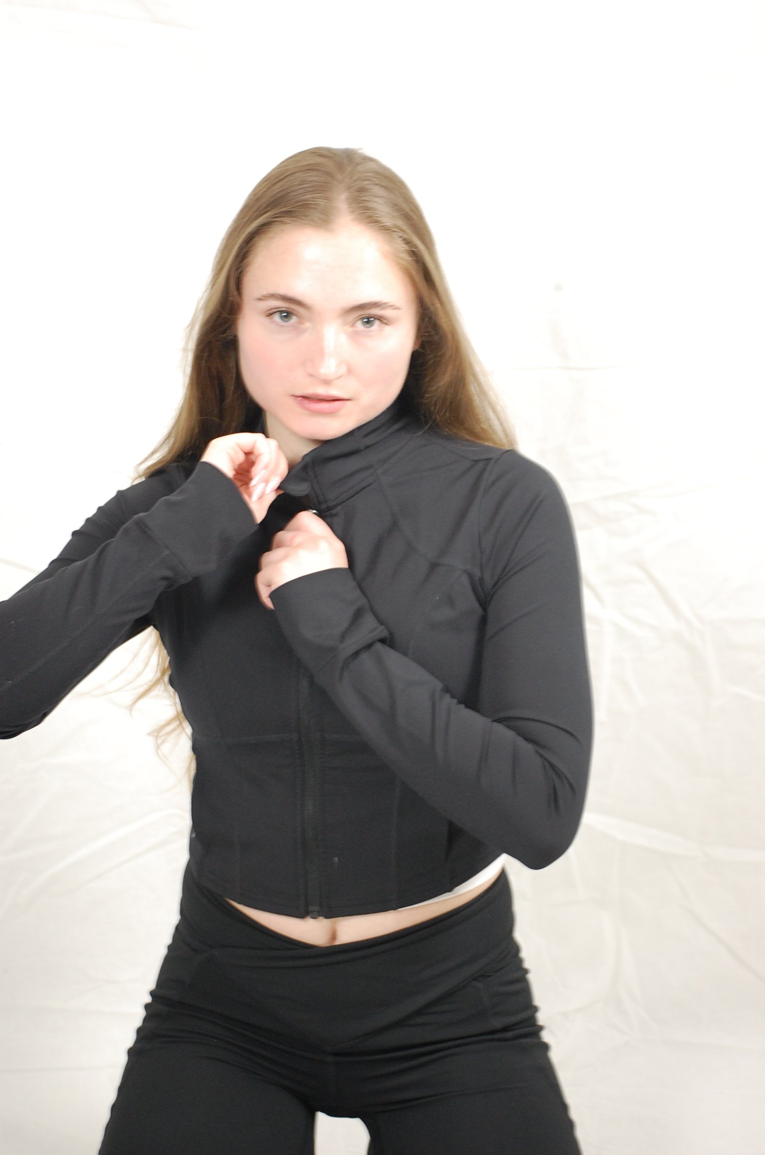 A young woman with long brown hair and fair skin wearing a black zip-up jacket and black pants, posing in front of a white background, with her left hand touching her jacket near her neck.