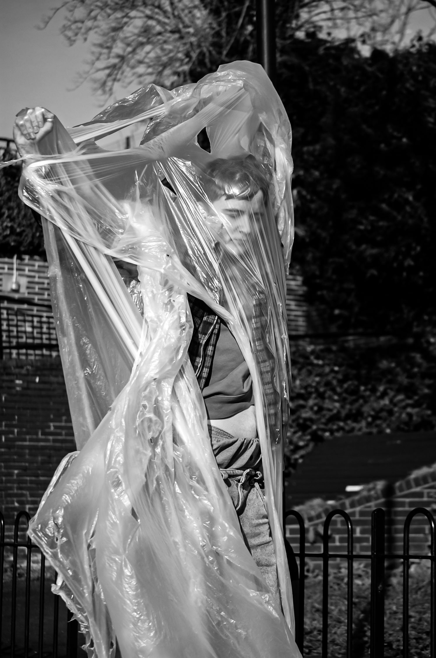 A young person with curly hair looking down, partially obscured by a large, transparent plastic bag, outdoors near a black metal fence and brick building.