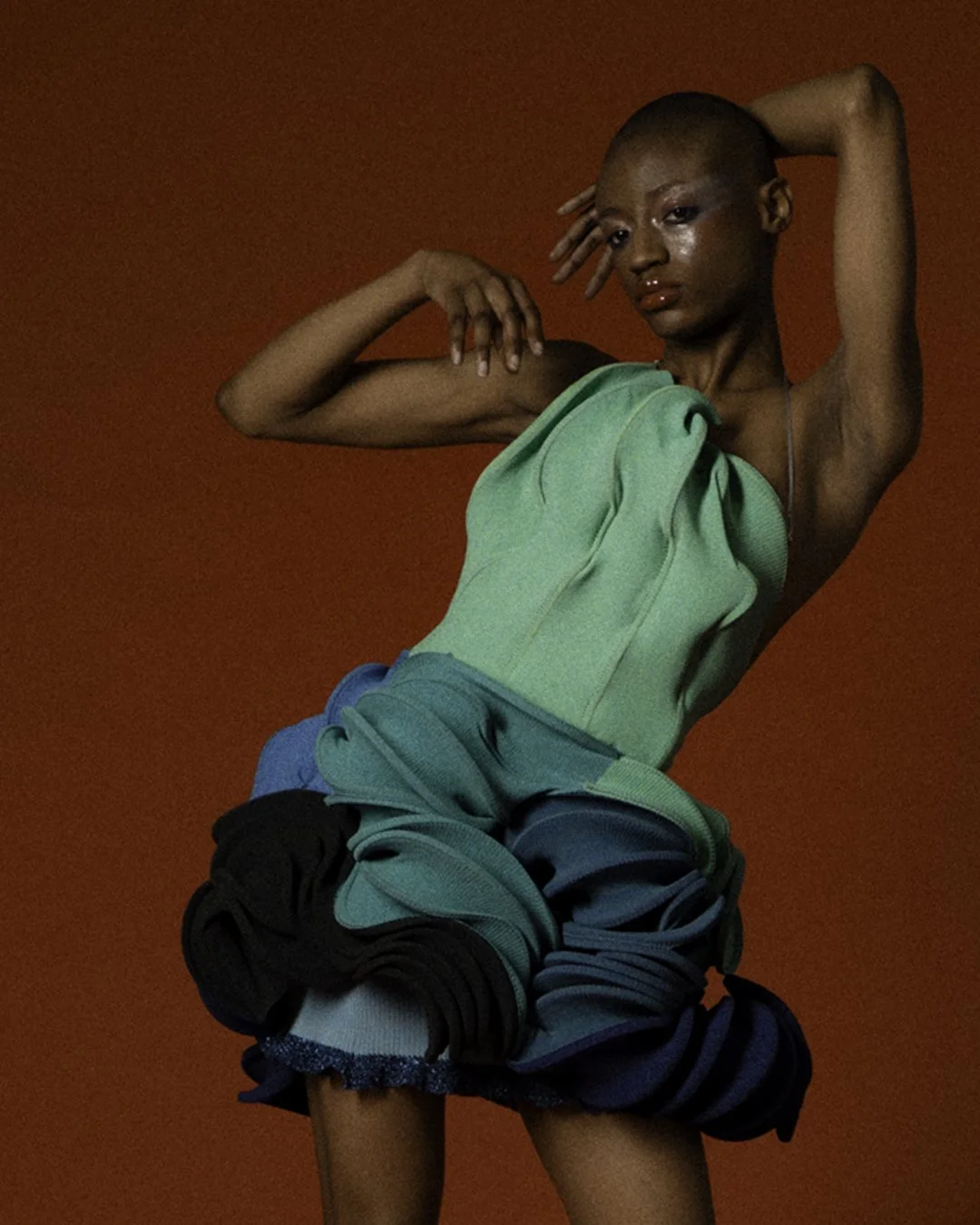 Fashion-forward woman with a shaved head and extended arm poses against a red background, wearing a vibrant green, layered, fabric dress and black layered skirt.