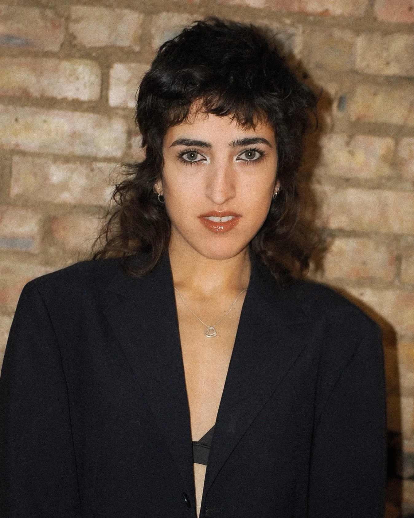 A woman with short, curly black hair and light-colored eyes wearing a black blazer, a silver necklace, and small earrings, standing in front of a brick wall.