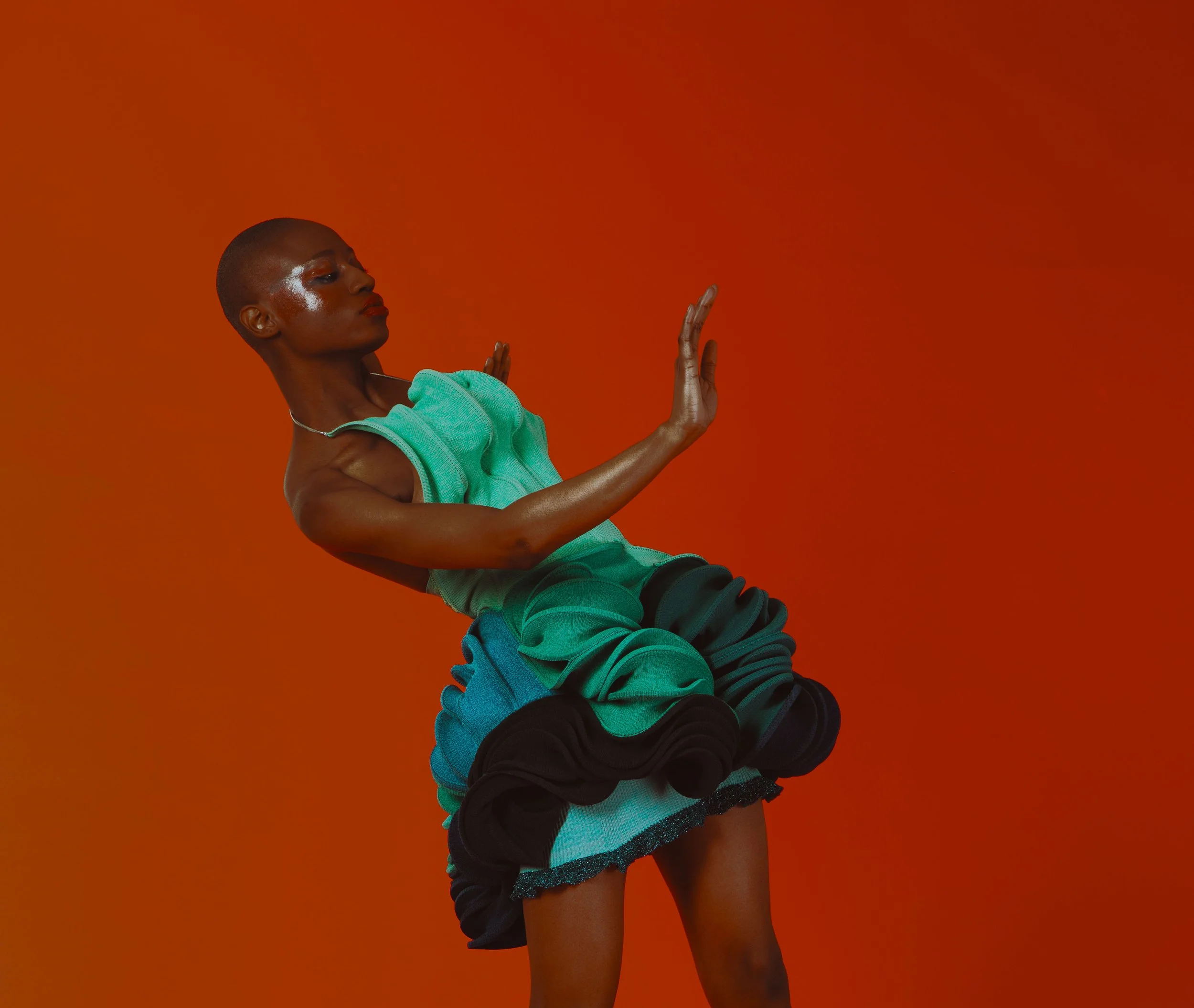 A person with a shaved head and dark skin wearing a turquoise top and a layered skirt, striking a dance pose against an orange background.